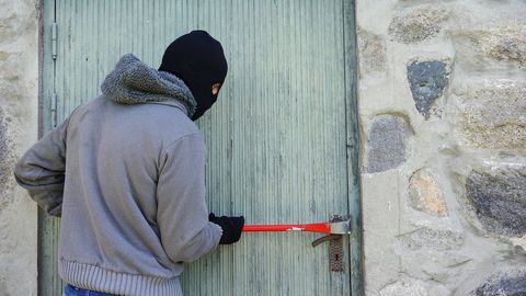 Auf dem Bild ist eine vermummte Person zu sehen, die mit einem roten Brecheisen versucht, eine Holztür aufzubrechen.