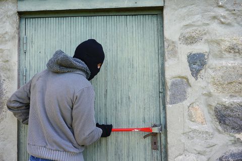 Auf dem Bild ist eine vermummte Person zu sehen, die mit einem roten Brecheisen versucht, eine Holztür aufzubrechen.